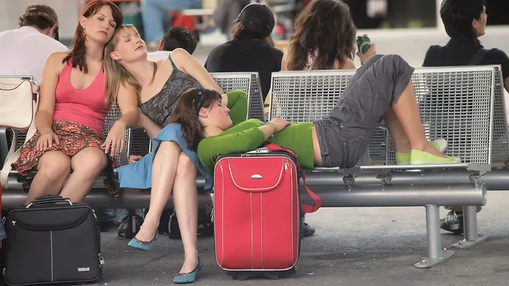 Man erkennt drei Frauen, die auf einer Wartebank am Flughafen sitzen. 