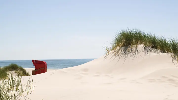 Au dem Foto ist eine Sanddüne zu sehen. In den Sanddünen steht ein roter Strandkorb. 
