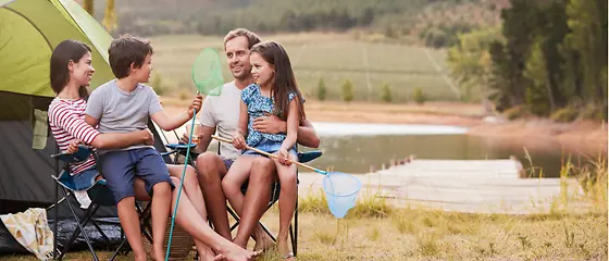 Familie beim Campen - Reiseschutz für den Camping-Urlaub Mutter, Vater und ihre Kinder sitzen vor einem Zelt. Im Hintergrund ist Wasser zu sehen.