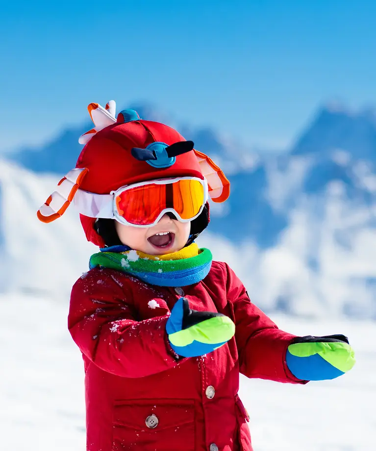 Ein Kind in einem roten Skianzug und rotem Skihelm. Im Hintergrund sind schneebedeckte Berge. 