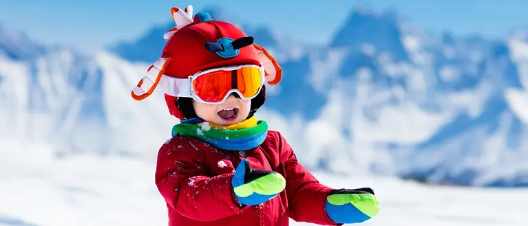 Ein Kind in einem roten Skianzug und rotem Skihelm. Im Hintergrund sind schneebedeckte Berge. 