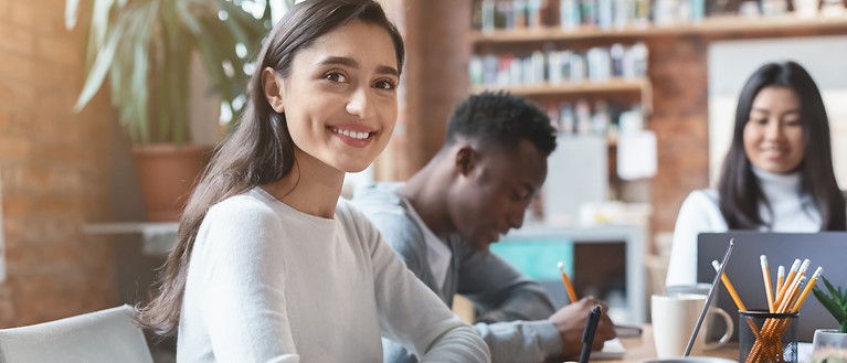 Drei Studenten sitzen an einem Tisch in einer Bibliothek und arbeiten. 