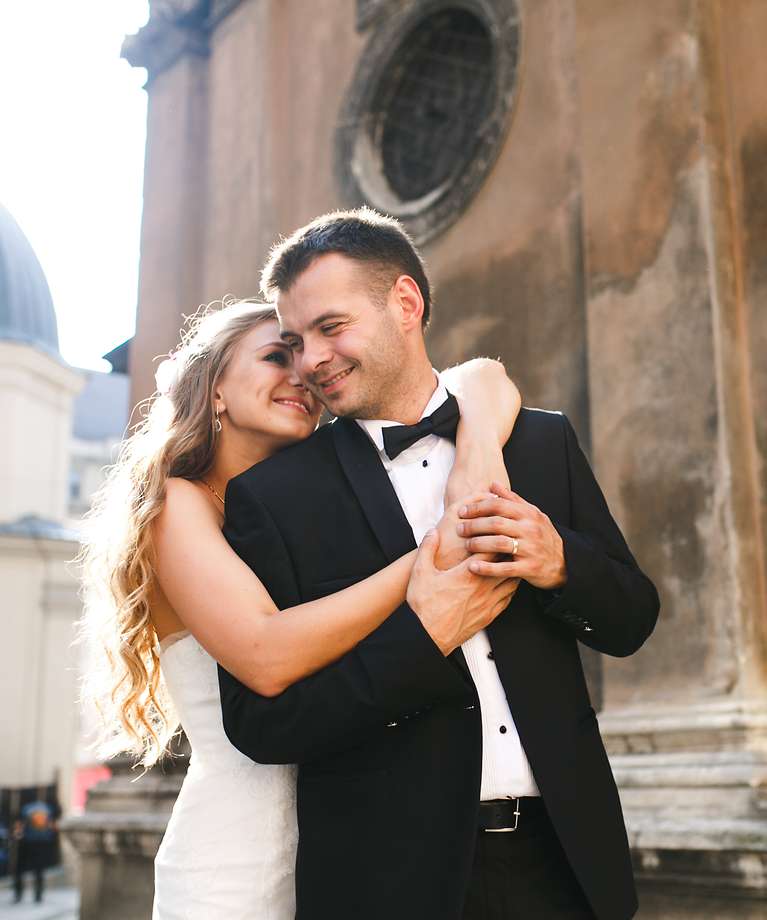 Eine Frau im Brautkleid und ein Mann im Anzug stehen vor der Kirche und umarmen sich. Beide lächeln sich an.