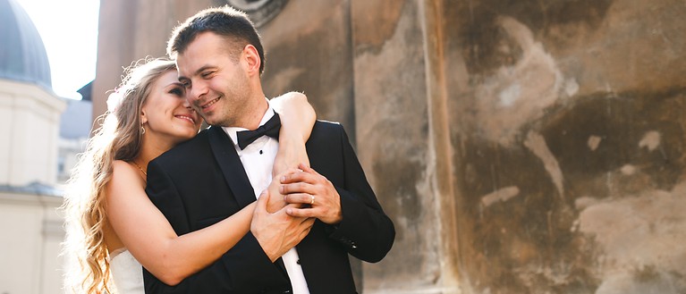 Eine Frau im Brautkleid und ein Mann im Anzug stehen vor der Kirche und umarmen sich. Beide lächeln sich an.