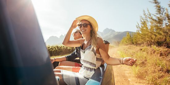 Eine Frau und ein Mann sitzen auf der Ladefläche eines Autos und genießen die Sonne. Im Hintergrund sind die Berge. 