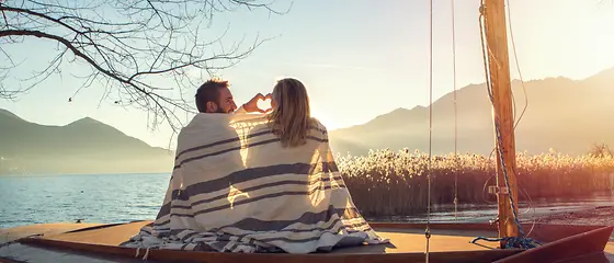 Pärchen am Wasser - Reiseschutz Gold Zwei Erwachsenen Menschen sitzen mit einer Decke umschlungen am Wasser. Im Hintergrund geht die Sonne unter.