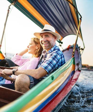 Ein Ehepaar sitzt in einem bunten Boot. Im Hintergrund kann man Häuser sehen.