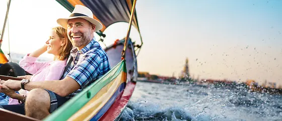 Ehepaar in einem Boot - Reiserücktrittsversicherung & Reiseabbruch-Versicherung Ein Ehepaar sitzt in einem bunten Boot. Im Hintergrund kann man Häuser sehen.