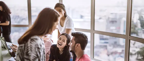 Junge Menschen am Lachen - Reiseversicherung für Schüler & Studenten Verschieden Personen sitzen an einem Tisch und lachen. Auf dem Tisch kann man Laptops sehen.