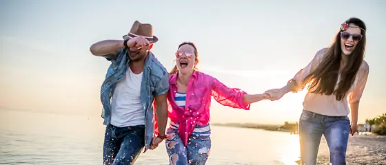 Freunde am Wasser - Last-Minute Reiseschutz Drei Freunde gehen am Wasser spazieren und tragen alle eine Sonnenbrille.