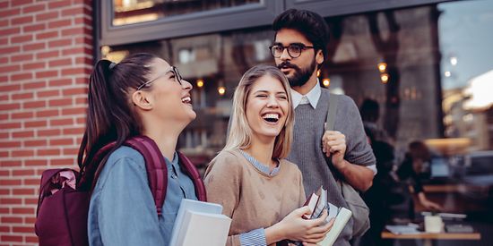 Drei Studenten mit Büchern in der Hand lachen. Im Hintergrund kann man ein Café sehen.