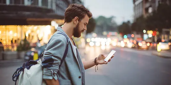 Ein Mann steht mit einem Handy in der Hand an einer Straße. Im Hintergrund sind Geschäfte zu sehen. 