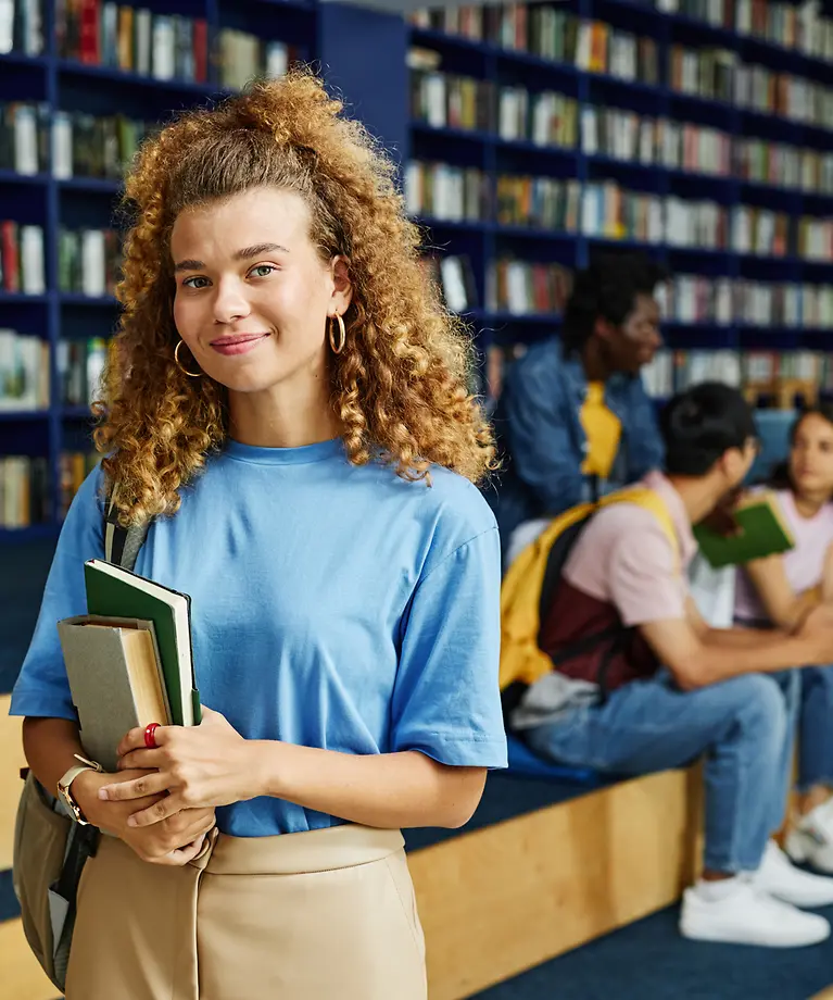Eine junge Studentin steht mit einem Buch in der Hand in einer Bibliothek.